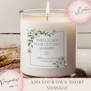 May include: A burning white candle in a clear glass jar. The label reads "Smells Like Your Getting Married" with floral details. The candle is a wedding or bridal shower gift, with space for a message.