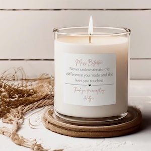 Op de afbeelding: Een brandende witte kaars in een doorzichtige glazen pot met een vierkant etiket met de tekst: Miss Betterton, Never underestimate the difference you made and the lives you touched. Thank you for everything, Holly x. De kaars staat op een houten onderzetter.