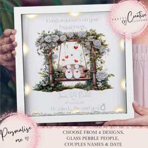 Op de afbeelding: Een wit frame met een verlichtingsfunctie, waarop een aquarel-illustratie te zien is van een stel dat op een schommel zit onder een bloemenboog. De tekst "Congratulations on your Engagement" staat bovenaan het frame. De namen "Jamie & Rachel" en de datum "18 February 2023" staan onder de illustratie. De tekst "He asked... She said yes!" staat onder de datum. Het frame is versierd met glitter en een klein ringpictogram.