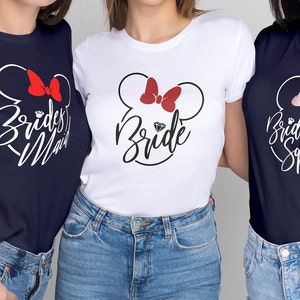 May include: Three navy blue t-shirts with white text and a white outline of Mickey Mouse ears with a red bow. The text on the shirts reads "Bride", "Bridesmaid", and "Bride Squad".