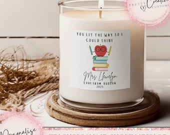 May include: A lit candle in a clear glass jar with a white label. The label features an apple with glasses, books, and the text "YOU LIT THE WAY SO I COULD SHINE" and "Mrs Hewelyn LOVE FROM HUDSON 2025". The candle sits on a wooden coaster.