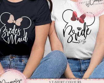 Puede incluir: Dos camisetas blancas con un diseño de orejas de Mickey Mouse negro. Una camiseta dice "Bride to Be" con un lazo rojo y la otra dice "Bridesmaid" con un lazo rosa.