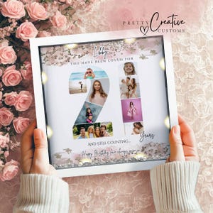 May include: A square white shadow box frame containing a photo collage arranged in the shape of the number 21. The design features the text Libby, You have been loved for 21 years, and still counting, with pink and gold glitter accents.