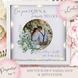 May include: A white framed wedding anniversary photo with the text "One year DOWN & Forever TO GO..." and "Anthony & Charlotte 21ST JUNE 2024". The photo features a couple inside a gold circle, surrounded by greenery and lights. The frame includes the text "Personalise me" and "ADD YOUR OWN NAMES, DATE & DESTINATION".