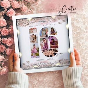 Può includere: Una cornice quadrata bianca tipo shadow box contenente un collage fotografico disposto a forma del numero 16. Il testo recita Amelia, You have been loved for 16 years, and still counting, Happy 1st Birthday love always, Nan xx. Logo Pretty Creative Customs.