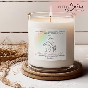 Puede incluir: Una vela en vaso de cristal transparente con cera blanca y una mecha encendida. La etiqueta muestra un dibujo lineal negro de Winnie the Pooh y Piglet, un degradado de arcoíris y el texto: It makes such a difference, said Pooh, to have someone who believes in you. Miss Armstrong, thank you for being such an important part of my story. From Lloyd.