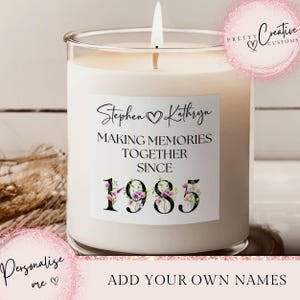 Puede incluir: Una vela blanca encendida en un tarro de cristal transparente. La vela tiene una etiqueta con los nombres "Stephen & Kathryn", el texto "MAKING MEMORIES TOGETHER SINCE 1985" y detalles florales. La vela está personalizada.