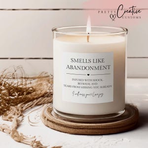 May include: A white wax candle in a clear glass jar with a lit wick. The label reads: SMELLS LIKE ABANDONMENT, INFUSED WITH SHOCK, BETRAYAL AND TEARS FROM MISSING YOU ALREADY, I will miss you!!! Love jag. The candle sits on a round wooden coaster.