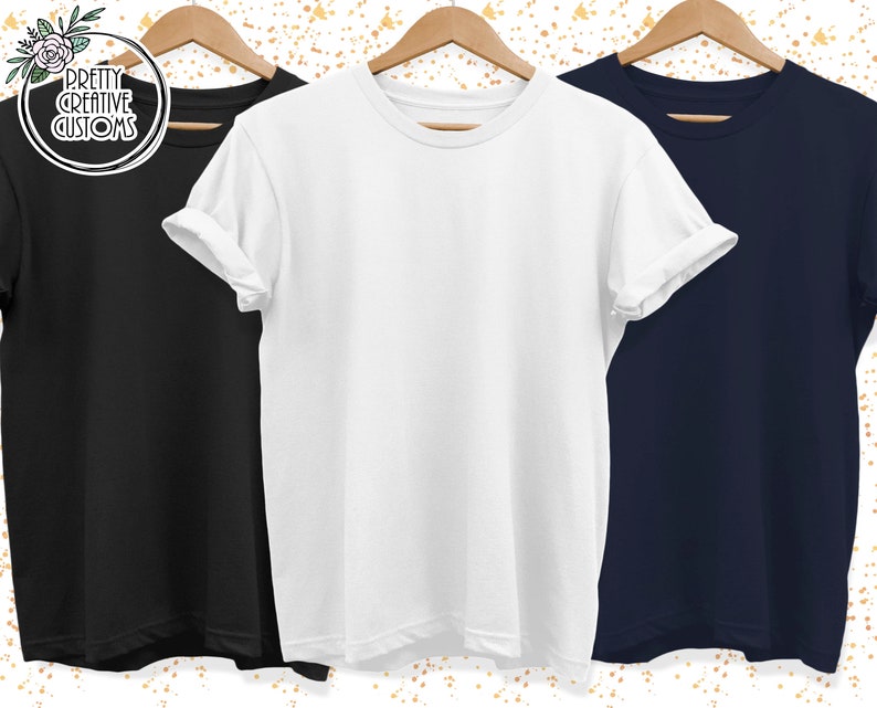 May include: Three blank t-shirts, one black, one white, and one navy blue, hanging on wooden hangers. The shirts are all plain and have no designs or text.
