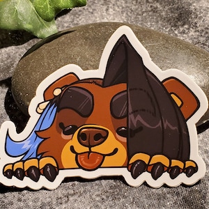 May include: A brown and black cartoon bear with a blue scarf peeking out from behind a black rock. The bear has a happy expression with its tongue sticking out.