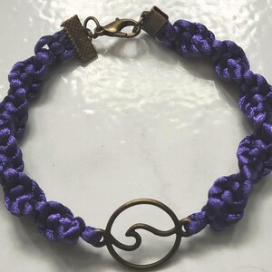 May include: A braided purple bracelet with a bronze-colored wave charm. The bracelet is made of woven cord and features a bronze clasp. The wave charm is a simple, open design. This handcrafted bracelet is a unique accessory.
