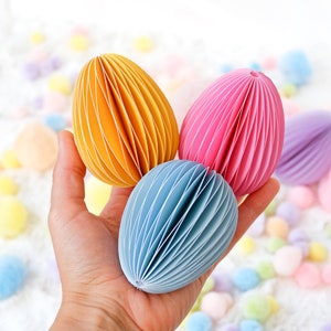 May include: A hand holds three colorful paper Easter eggs in yellow, pink, and blue. The eggs have a layered, accordion-like design. Pastel-colored pom-poms are scattered in the background, creating a festive, spring-like scene.