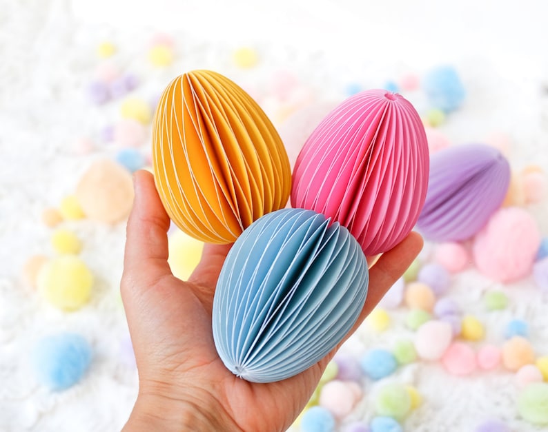May include: A hand holds three colorful paper Easter eggs in yellow, pink, and blue. The eggs have a layered, accordion-like design. Pastel-colored pom-poms are scattered in the background, creating a festive, spring-like scene.