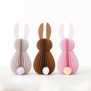 May include: Three paper Easter bunny decorations in neutral colors. The bunnies are made of layered paper with a pom-pom tail. One is light gray, one is brown, and one is pink. The ears are a lighter shade of the body color.