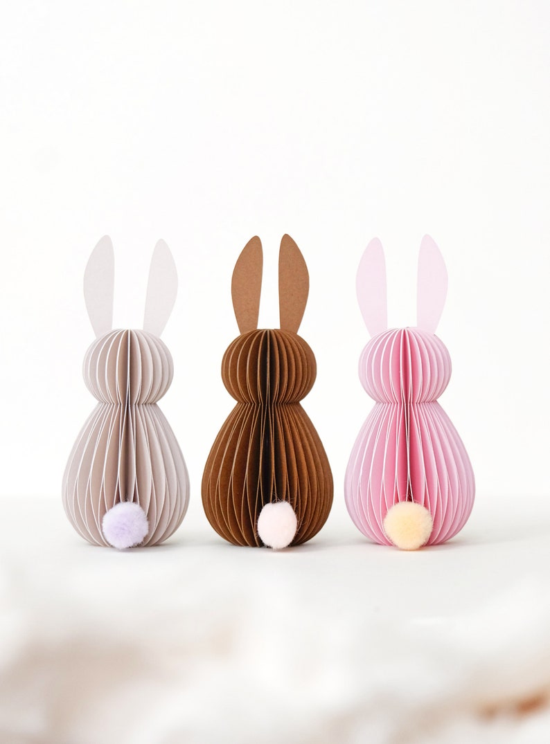 May include: Three paper Easter bunny decorations in neutral colors. The bunnies are made of layered paper with a pom-pom tail. One is light gray, one is brown, and one is pink. The ears are a lighter shade of the body color.