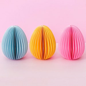 May include: Three paper egg-shaped decorations in blue, yellow, and pink. Each egg has a ribbed design and sits on a pink surface against a pink background. The eggs are evenly spaced and create a colorful, decorative display.