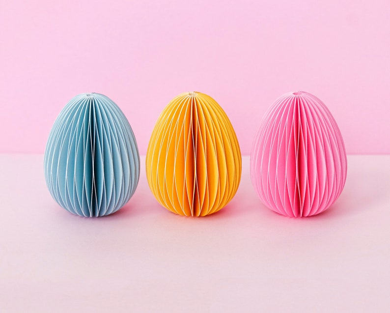 May include: Three paper egg-shaped decorations in blue, yellow, and pink. Each egg has a ribbed design and sits on a pink surface against a pink background. The eggs are evenly spaced and create a colorful, decorative display.