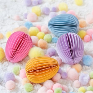 May include: An assortment of colorful, egg-shaped paper notepads in pink, blue, purple, and yellow. The notepads are surrounded by pastel-colored pom-poms on a white, textured surface. The notepads have a unique, layered design.