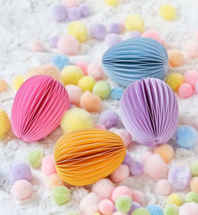 May include: An assortment of colorful, egg-shaped paper notepads in pink, blue, purple, and yellow. The notepads are surrounded by pastel-colored pom-poms on a white, textured surface. The notepads have a unique, layered design.