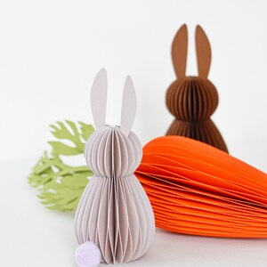 May include: Paper Easter decorations featuring a light gray bunny with a lavender pom-pom tail, a brown bunny, and an orange carrot. A green paper cutout of a carrot top is also present. The bunny figures are made of layered paper.