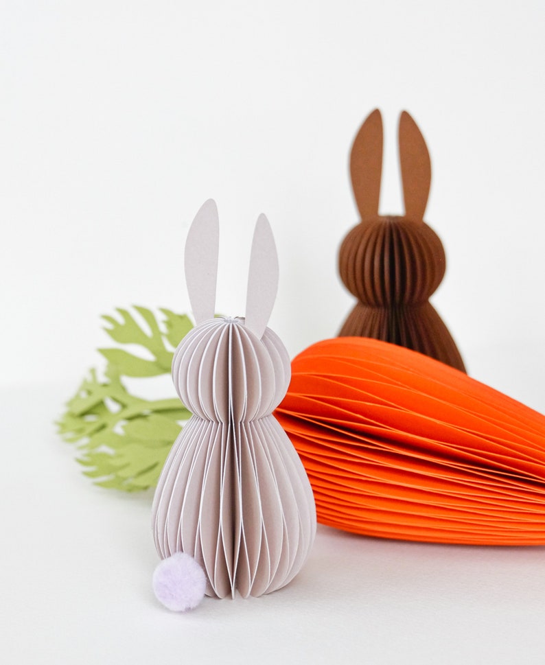 May include: Paper Easter decorations featuring a light gray bunny with a lavender pom-pom tail, a brown bunny, and an orange carrot. A green paper cutout of a carrot top is also present. The bunny figures are made of layered paper.