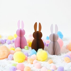 May include: Three paper Easter bunny decorations in pink, brown, and beige, with matching ear colors. The bunnies are surrounded by colorful pom-poms on a white, fluffy surface, creating a festive spring scene.