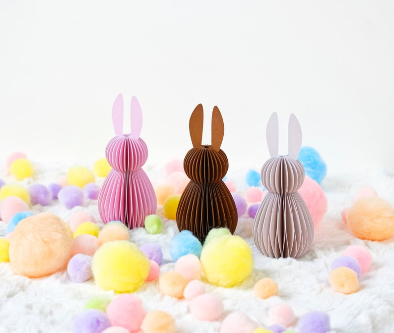 May include: Three paper Easter bunny decorations in pink, brown, and beige, with matching ear colors. The bunnies are surrounded by colorful pom-poms on a white, fluffy surface, creating a festive spring scene.