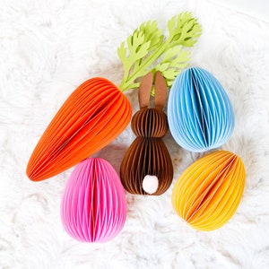 May include: An Easter-themed arrangement featuring a paper carrot, a brown paper bunny, and three paper eggs in pink, blue, and yellow. The carrot and eggs have a ribbed design, and the bunny has a white tail. Green paper foliage is also included.