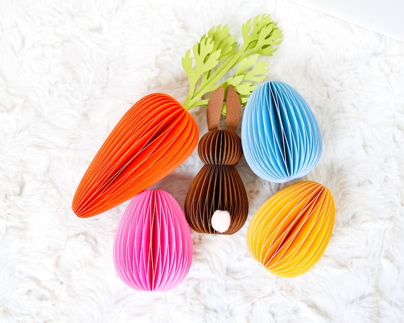 May include: An Easter-themed arrangement featuring a paper carrot, a brown paper bunny, and three paper eggs in pink, blue, and yellow. The carrot and eggs have a ribbed design, and the bunny has a white tail. Green paper foliage is also included.