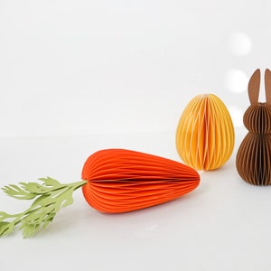 May include: An Easter-themed paper craft arrangement featuring a large orange carrot with green leaves, a yellow egg, and a brown bunny. The objects are made of folded paper, creating a textured effect. The background is plain white.