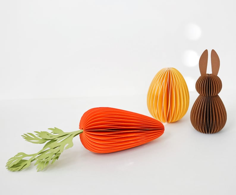 May include: An Easter-themed paper craft arrangement featuring a large orange carrot with green leaves, a yellow egg, and a brown bunny. The objects are made of folded paper, creating a textured effect. The background is plain white.