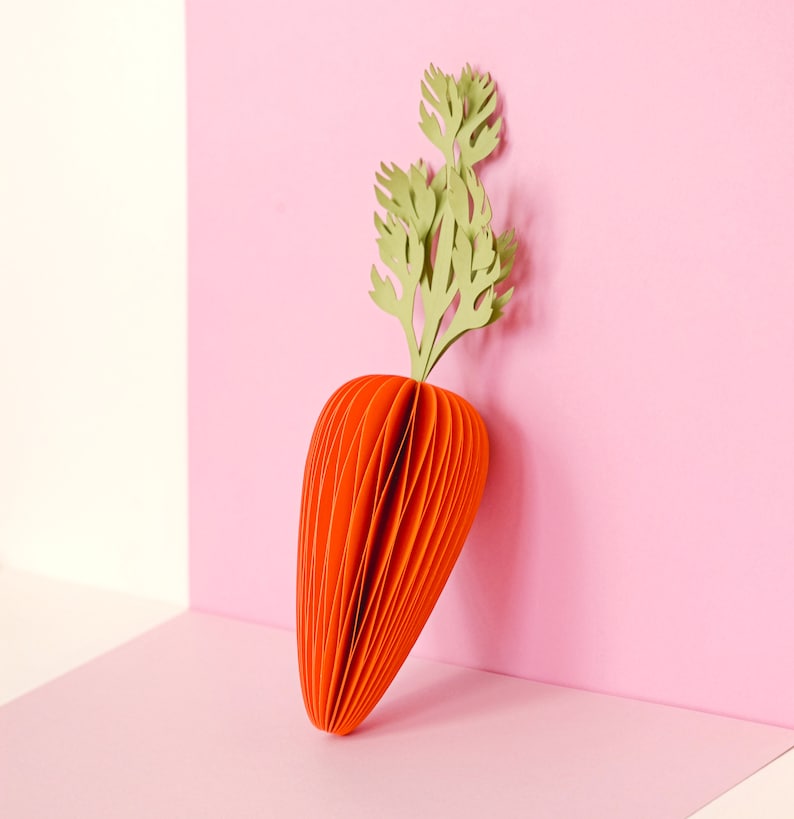May include: A paper carrot sculpture with an orange body and green leafy top. The carrot is made of layered paper, creating a textured effect. The background is a two-tone pink and white.