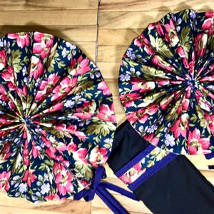 May include: Two fabric fan-shaped decorations with a floral print in shades of pink, green, and purple on a dark blue background. The fans are tied together with purple ribbon.