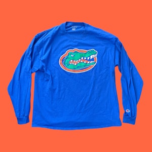 May include: A blue long-sleeve t-shirt with a green alligator head graphic and the word "Champion" in white on the right sleeve.