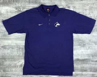 Op de afbeelding: Paars poloshirt met een wit Nike swoosh logo en een wit husky logo op de voorkant. Het shirt heeft een knoopsluiting en korte mouwen.
