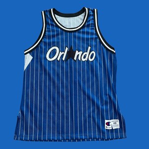 May include: A blue and white striped basketball jersey with the word "Orlando" and a star on the front. The jersey has a black trim and a Champion logo on the bottom right corner. The jersey is size 48.