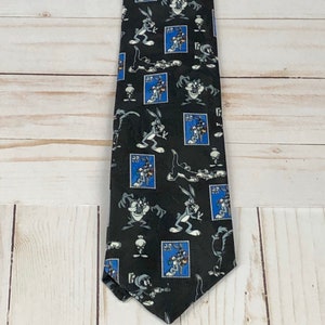 May include: A black tie with a repeating pattern of Bugs Bunny and Tweety Bird cartoon characters on blue postage stamps. The tie is made of a silky material.
