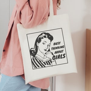 May include: A white tote bag with a black and white illustration of a woman with the text "Busy thinking about girls".