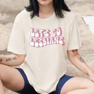 May include: Cream-colored t-shirt with the text "LET'S GO LESBIANS" in a retro, bubble-letter style. The text is in white with a pink and orange outline. The shirt is paired with navy blue shorts.
