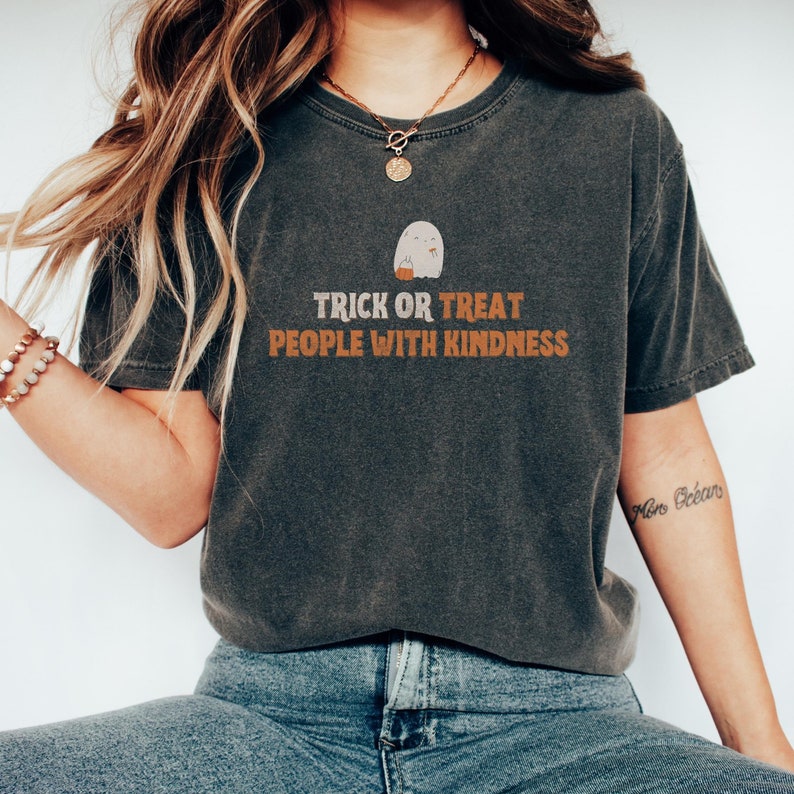 May include: A dark gray t-shirt with a white ghost holding a pumpkin bucket. The text "Trick or Treat People with Kindness" is printed in orange on the shirt.