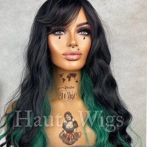 May include: A long, wavy black and green wig with a side part and bangs. The wig is styled with loose waves and has a green streak on one side. The wig is on a mannequin head with a tattoo of a world map and the words "Haute Wig" on the neck.