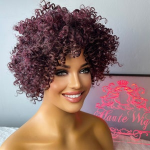Peut inclure: Une perruque courte et bouclée de couleur bordeaux foncé. La perruque est coiffée en afro volumineux avec des boucles lâches. La perruque est sur une tête de mannequin avec un maquillage neutre.
