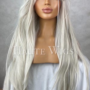 Könnte beinhalten: Lange, silberblonde Perücke mit einem seitlich geschwungenen Pony. Die Perücke ist auf einem Schaufensterkopf gestylt, mit dem Text "HAUTE WIGS" auf der Vorderseite. Das Haar ist lang und fließend, mit einer natürlich aussehenden Textur.