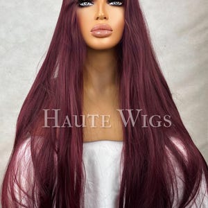 May include: A long, burgundy wig with a fringe and long layers. The wig is styled on a mannequin head with the text "Haute Wigs" in the centre. The wig has a natural-looking hairline and is made of synthetic fibres.
