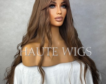 May include: A long, wavy, light brown wig with bangs. The wig is styled with loose waves and a side part. The wig is on a mannequin head. The text "HAUTE WIGS" is visible on the mannequin's chest.