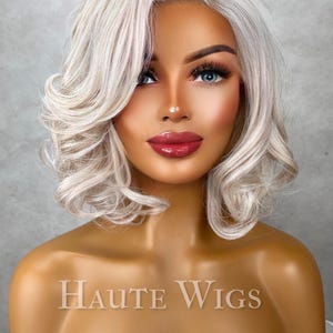 Könnte beinhalten: Ein Schaufensterpuppenkopf mit einer kurzen, gewellten, aschblonden Perücke. Die Perücke hat einen Seitenscheitel und weiche Locken. Die Schaufensterpuppe hat ein komplettes Make-up, einschließlich rotem Lippenstift und blauen Augen. Der Text "Haute Wigs" ist sichtbar.