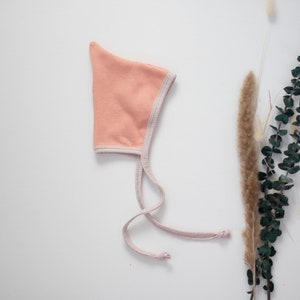 May include: A peach-colored baby bonnet with a white trim and ties. The bonnet is made of soft fabric and is perfect for keeping a baby's head warm.