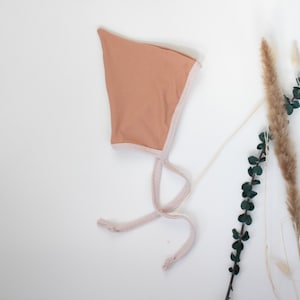 May include: A peach-colored baby bonnet with a white trim and ties. The bonnet is on a white background with dried flowers and greenery.