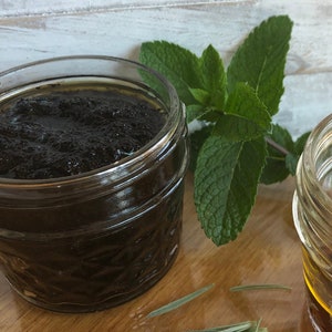 May include: Two glass jars with lids, one filled with a dark brown liquid and the other with a light brown liquid. A sprig of green mint and a sprig of lavender are on a wooden surface.