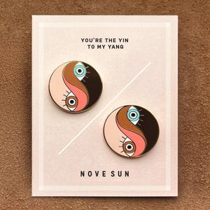 May include: Two enamel pins in the shape of a yin and yang symbol, each with an eye design. The pins are on a pink card with the text "YOU'RE THE YIN TO MY YANG" and "NOVE SUN". The pins feature brown, pink, and blue colors.
